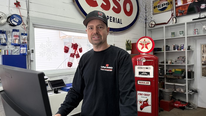 Tony Mercier est derrière la caisse enregistreuse du garage. Derrière lui, on remarque une grande affiche Esso et une pompe à essence antique aux couleurs de Texaco.