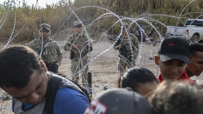 Migrants walk in front of National Guard soldiers standing guard behind a barbed wire fence.