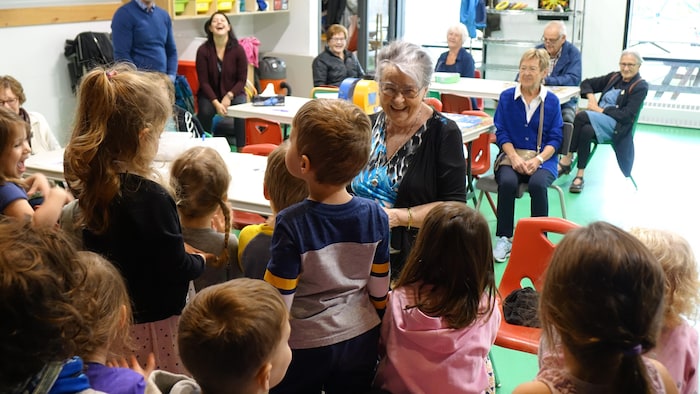 Une dame aux cheveux blancs avec des lunettes raconte l'histoire, entourée d'enfants.