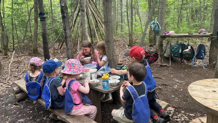 Des enfants mangent une collation sur des tables et des bancs en bois, au milieu d'une forêt, sous la supervision d'une éducatrice.