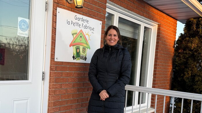 Une femme en manteau pose en souriant sur le balcon d'un bâtiment. Une affiche portant le nom d'une garderie est visible sur la façade.