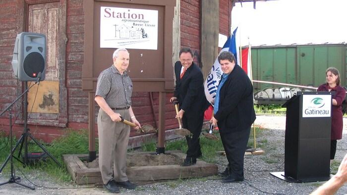 Trois personnes soulèvent de la terre avec une pelle devant l'ancienne gare de Masson-Angers le 27 juin 2005. De gauche à droite : Rolland Pagé, membre du comité de relance, Yves Ducharme, maire de Gatineau, et Patrick Duguay, membre du comité de relance.