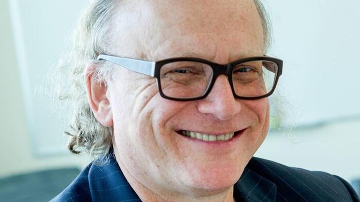 Un homme portant des lunettes sourit en fixant l'appareil-photo.