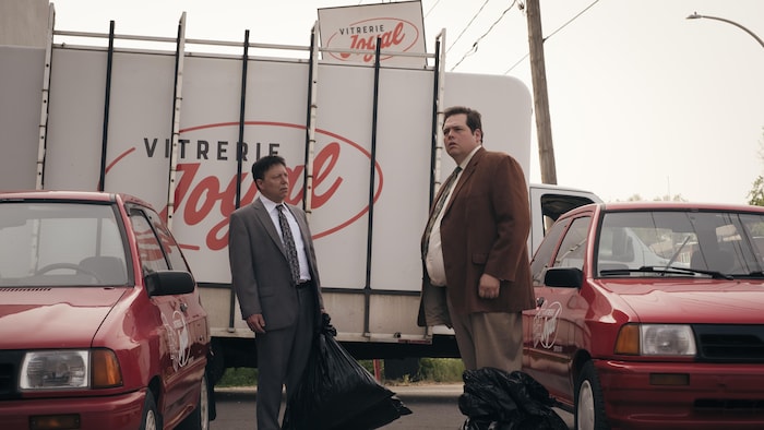 Deux hommes tiennent des sacs de vidange devant leur voiture rouge, devant des affiches de Vitrerie Joyal. 