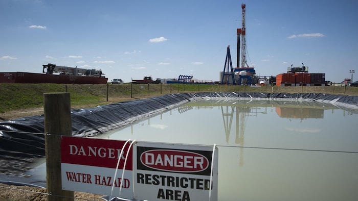 Un bassin de rétention d'eau bordé d'une clôture portant des panneaux de danger se trouve devant un site industriel.