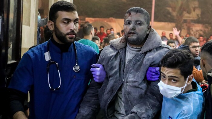 Un homme couvert de poussière et avec une blessure à la tête est soutenu par deux médecins. 