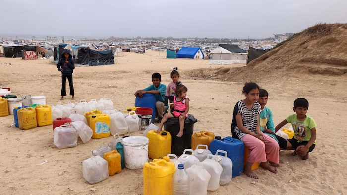 Des enfants sont assis sur des récipients destinés à contenir de l'eau, devant une multitude de tentes.