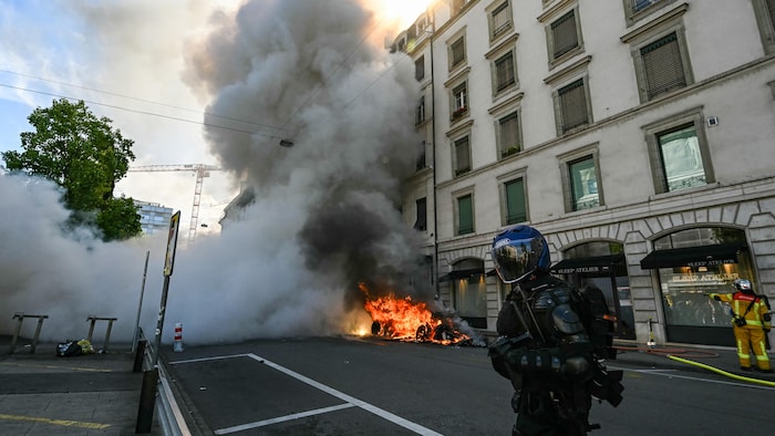 Un policier suisse en tenue antiémeute se tient à côté d'une Tesla en feu lors de la manifestation contre le G7 à Genève, le 14&nbsp;juin&nbsp;2026. 