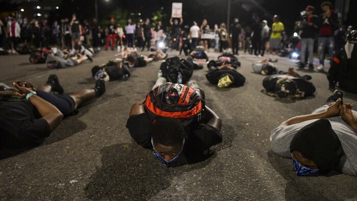 Plusieurs personnes sont couchés au milieu d'une rue, le visage vers le sol.