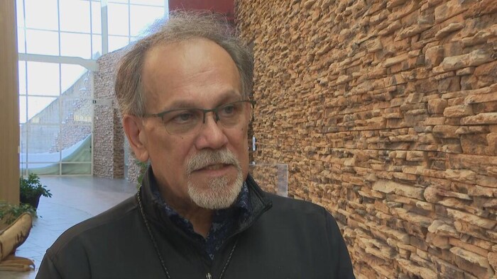 Un homme à l'intérieur d'un édifice, près d'un mur de briques, en train de parler.