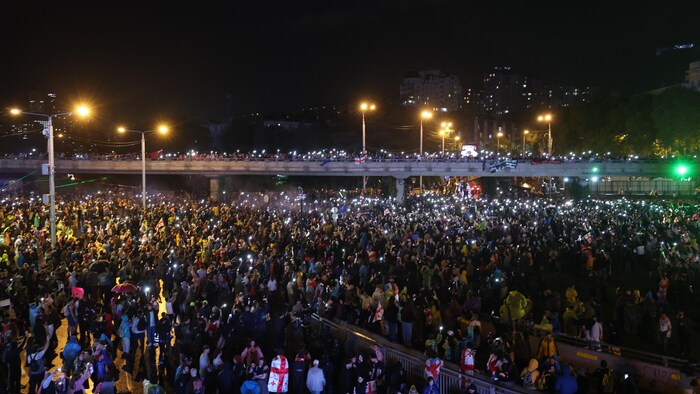 Des milliers de manifestants sont dans les rues et sur un pont durant le soir et arborent des drapeaux de la Géorgie. 