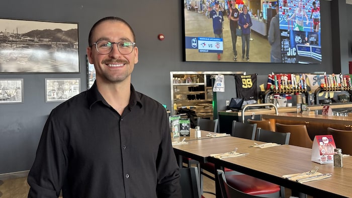 Un homme souriant à lunettes et moustache, vêtu d'une chemise noire, pose dans un restaurant de type bar sportif. L'arrière-plan montre de grands écrans diffusant du sport, des tables dressées et un comptoir de bar avec plusieurs pompes à bière.