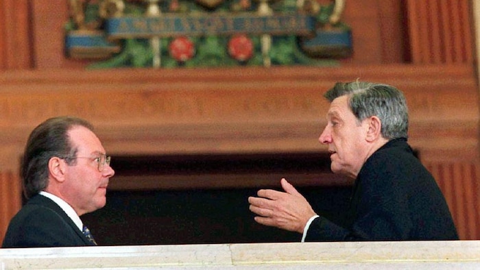 Sous les imposantes armoiries du Sénat du Canada, Michel Bastarache et Gérard La Forest apparaissent de profil. Gérard La Forest a la main tendue devant lui et parle.