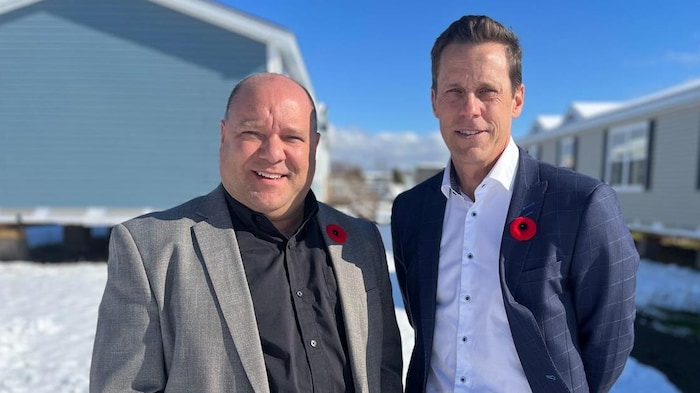 Deux hommes en veston posent pour une photo, debout dans la neige entre des maisons mobiles.