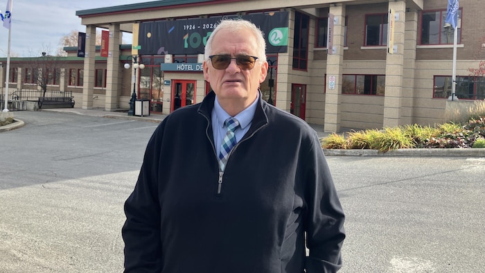 Gilles Chapadeau, lunettes de soleil sur les yeux, fixe la caméra en posant devant l'hôtel de ville de Rouyn-Noranda.