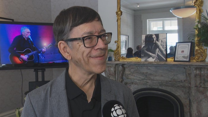 Dans une pièce où des photos et vidéos de Jacques Michel sont présentées, le chanteur Gilles Valiquette sourit, pendant une entrevue avec Radio-Canada.