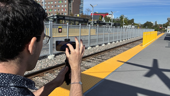 Gio Petti manipule son cellulaire attaché à un trépied sur le quai de l'O-Train.