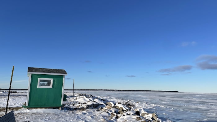 Une cabane de bois se dresse sur la berge englacée du fleuve. Au loin, l'Île Saint-Barnabé.