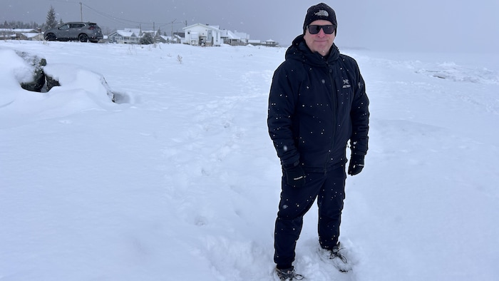 Dominique Bérubé debout près d'un quartier résidentiel et d'une baie gelée. Il porte une tuque et un manteau d'hiver. Le quartier et la baie sont enneigés.