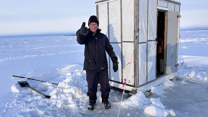 Alyre Hébert debout près de la cabane à éperlan sur la glace.