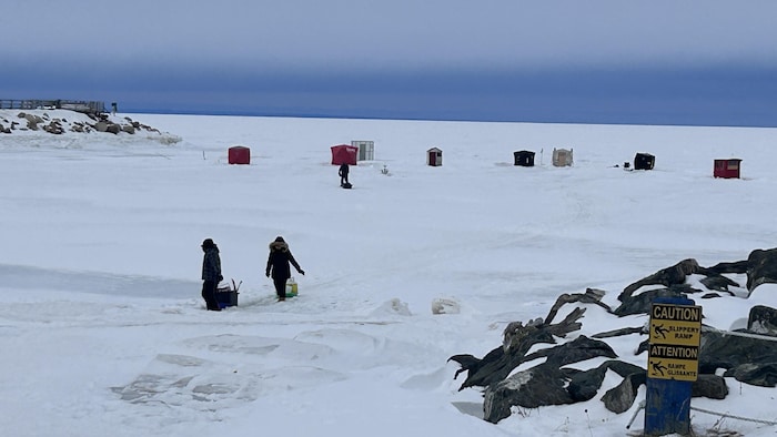 Des cabanes à éperlan sur la baie gelée. Deux pêcheurs marchent sur la glace.