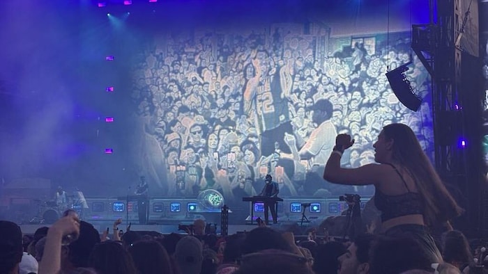 Une foule regarde une scène avec des musiciens et un écran géant montrant son chanteur parmi les festivaliers.