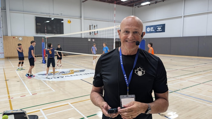 Glenn Hoag équipé d'un micro-casque lors d'une formation pratique en gymnase avec de jeunes volleyeurs en arrière-plan.