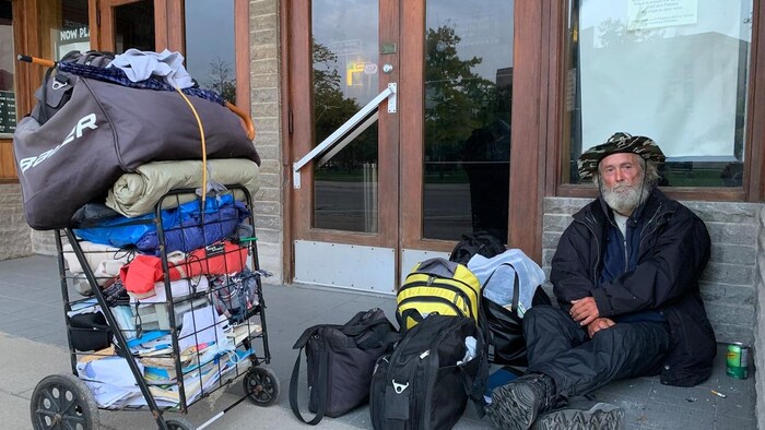 A man is sitting on the ground, by the entrance of a business. He is surrounded by bags and a trolley carrying his belongings.