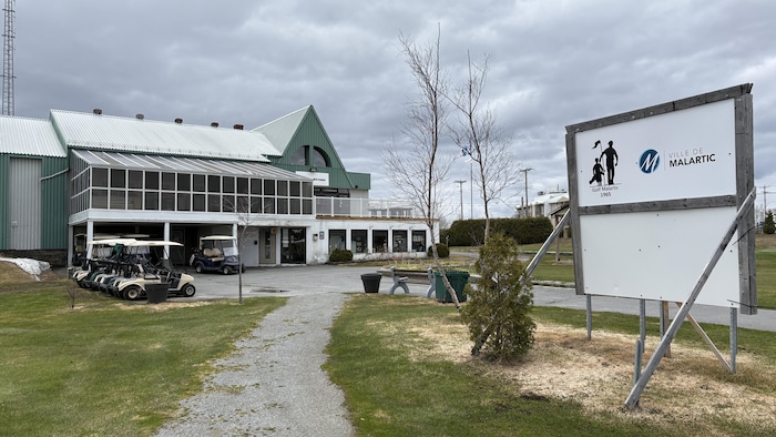 Des voiturettes électriques stationnées près du chalet du Club de golf de Malartic et à l'avant-plan, une affiche de la Ville de Malartic et du Golf Malartic.