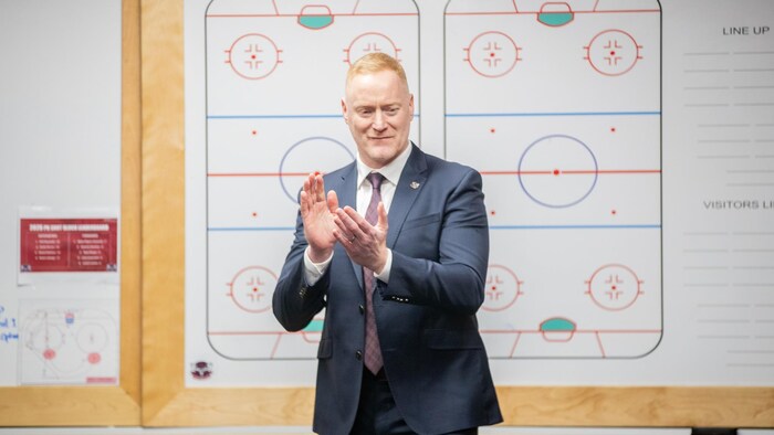 Un entraîneur en complet et cravate applaudit, debout devant un tableau avec des illustrations de patinoires de hockey.