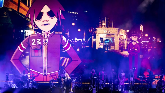 Un personnage animé est projeté derrière une scène où est installé un groupe de musique. 