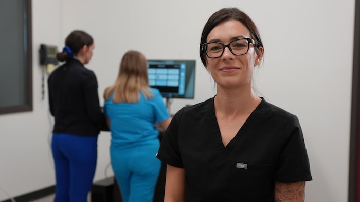 L'enseignante est au premier program  et regarde l'objectif, en arrière ses étudiante travaillent sur un ordinateur.