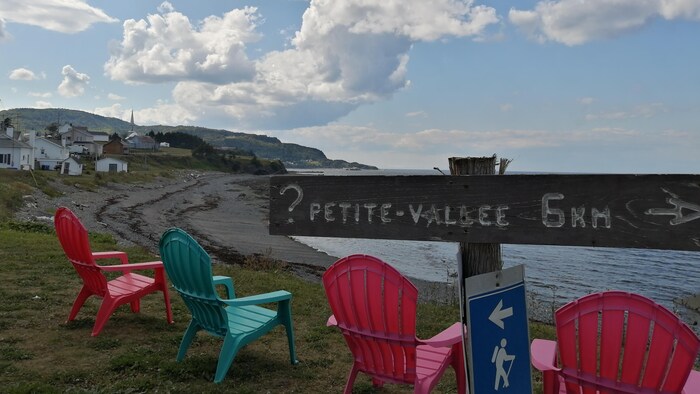 Une pancarte sur laquelle on peut lire : « Petite-Vallée, 5 km » devant des chaises installées face au fleuve.