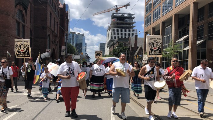 Des manifestants autochtones marchent dans la rue à Toronto avec des pancartes et des tambourins.