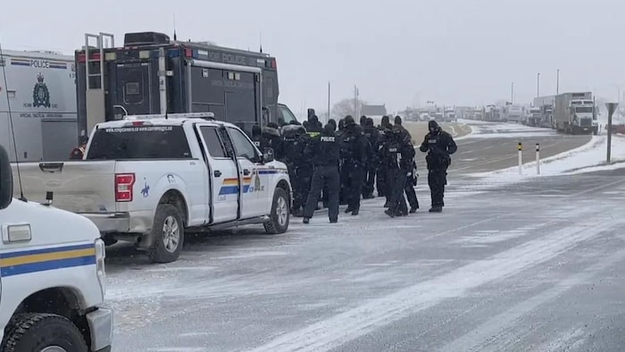 Une quinzaine d'agents de la G R C et des voitures de police se trouvent sur une route enneigée. Au loin, il y a une longue file de camions.