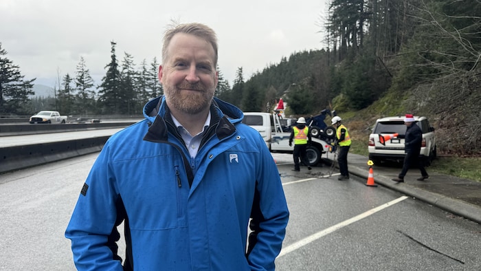 Greg Harper est sur le côté de l'autoroute. Derrière lui, un véhicule et un véhicule-remorque avec deux hommes vêtus d'habits de construction. Un autre homme a un cône de route dans une main.
