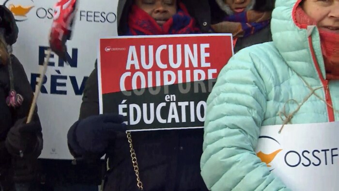 Des enseignants tiennent des pancartes, y compris une avec le message « aucune coupure en éducation ».