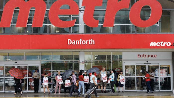 Des gens portent des pancartes où l'on peut lire les mots « En grève » devant les portes coulissantes d'un magasin Metro.