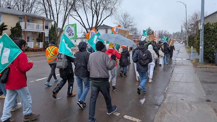 Une foule de manifestants marche sur une route mouillée bordée de maisons. Ils portent des vêtements d'hiver et agitent divers drapeaux colorés (verts, oranges, bleus). Un participant au centre tient un parapluie gris sous un ciel couvert et neigeux.