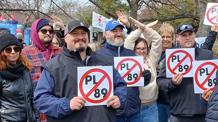 Un groupe de personnes proteste en croisant les bras et en tenant des pancartes qui portent un symbole d'abolition du projet de loi 89.