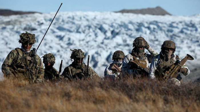 Six soldats en uniforme, armés, surgissent devant un haut monticule de neige dans un environnement arctique.
