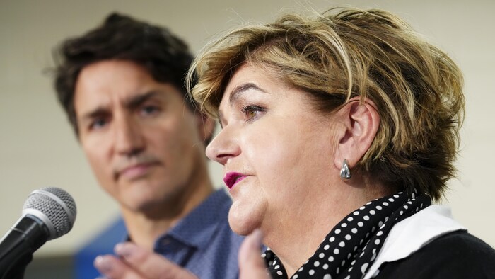 Le profil gauche de Gudie Hutchings, qui parle dans un micro en bougeant les mains, pendant que Justin Trudeau, derrière elle, la regarde.