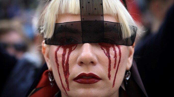 Une femme dont les yeux sont cachés par un ruban noir arbore un maquillage exprimant des larmes de sang coulant de ses yeux.