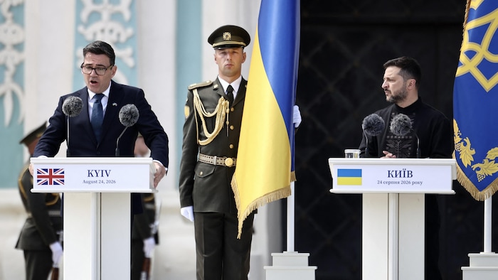 Andy Burnham et Volodymyr Zelensky derrière des podiums, alors qu'un soldat ukrainien tient un drapeau de son pays derrière eux.