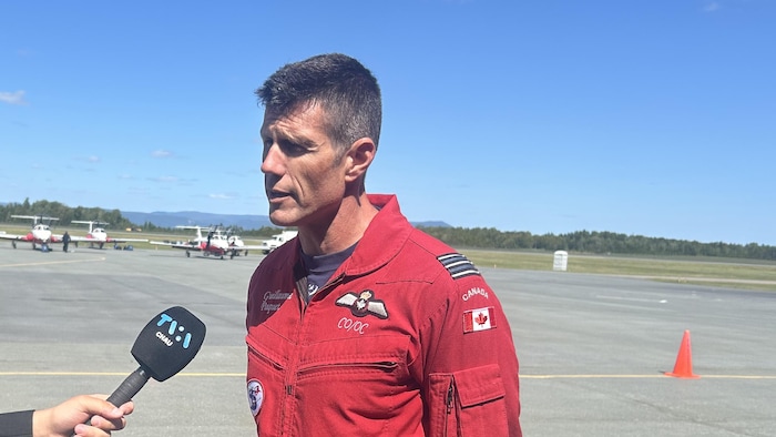Guillaume Paquet en uniforme des Snowbirds répond aux questions des journalistes sur le tarmac de l'aéroport de Charlo, des avions des Snowbirds derrière lui.