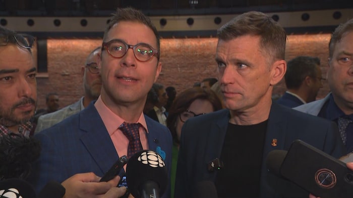 Deux hommes donnent un point de presse entouré de plusieurs personnes.