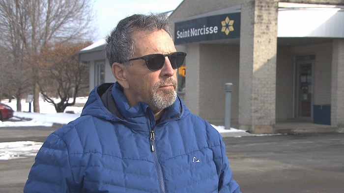 Un homme portant des lunettes fumés et un manteau d'hiver est debout devant le bureau municipal de Saint-Narcisse.
