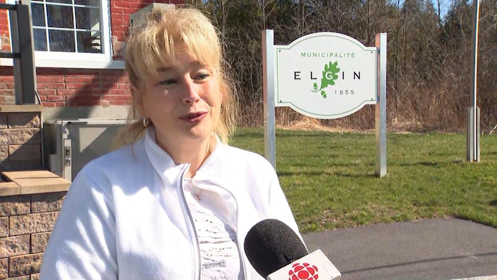 Guylaine Carrière est interviewée par une journaliste de Radio-Canada devant une pancarte affichant : Municipalité Elgin, 1855.