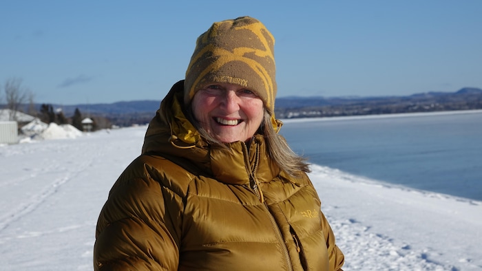 Une femme souriante avec une tuque regarde l'objectif, des mèches de cheveux sortent de son manteau d'hiver, devant la mer, à Maria.