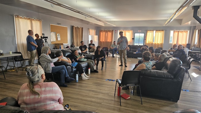 Une vingtaine de personnes sont assises dans une salle, et un individu se tient au centre, un microphone à la main. 